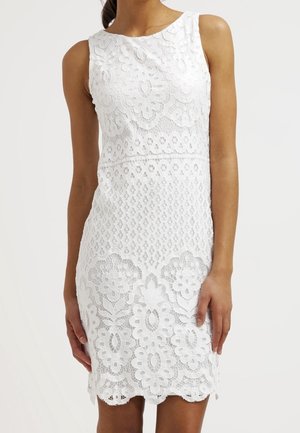 Femme portant une robe en dentelle blanche sans manches avec des motifs floraux et géométriques, debout les bras détendus le long du corps.
