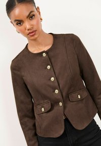 Veste en suede marron avec un col rond, fermetures à boutons dorés et deux poches avant, présentant un design ajusté et une texture lisse.