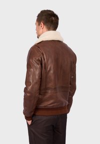 Hnědá kožená bunda s beránkovým límcem, žebrovaným pasem a detailním zdobením na ramenech. Hladká textura s bohatou, teplou barvou.