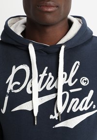 Hombre que lleva una sudadera azul marino con cordones blancos y texto en blanco parcialmente visible en el pecho.