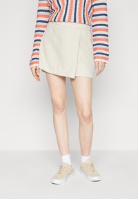 Jupe portefeuille beige clair, coupe droite et texture lisse, associée à un pull rayé coloré et des baskets beiges.