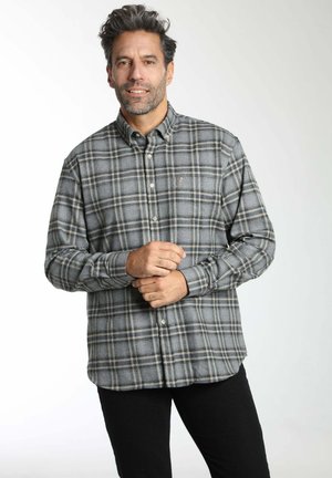 Homme aux cheveux gris portant une chemise à carreaux gris et noir boutonnée, debout les mains jointes, devant un fond clair uni.