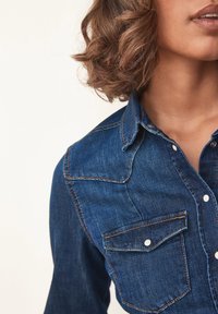 Cămașă din denim de culoare albastru închis, cu un design ajustat, clapetă cu nasturi, două buzunare pe piept și detalii de cusătură contrastante.