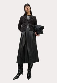 Un cardigan noir sur un top rayé, associé à une jupe plissée en cuir noir et des bottes à bout pointu noires et brillantes. Tenant un sac noir texturé.