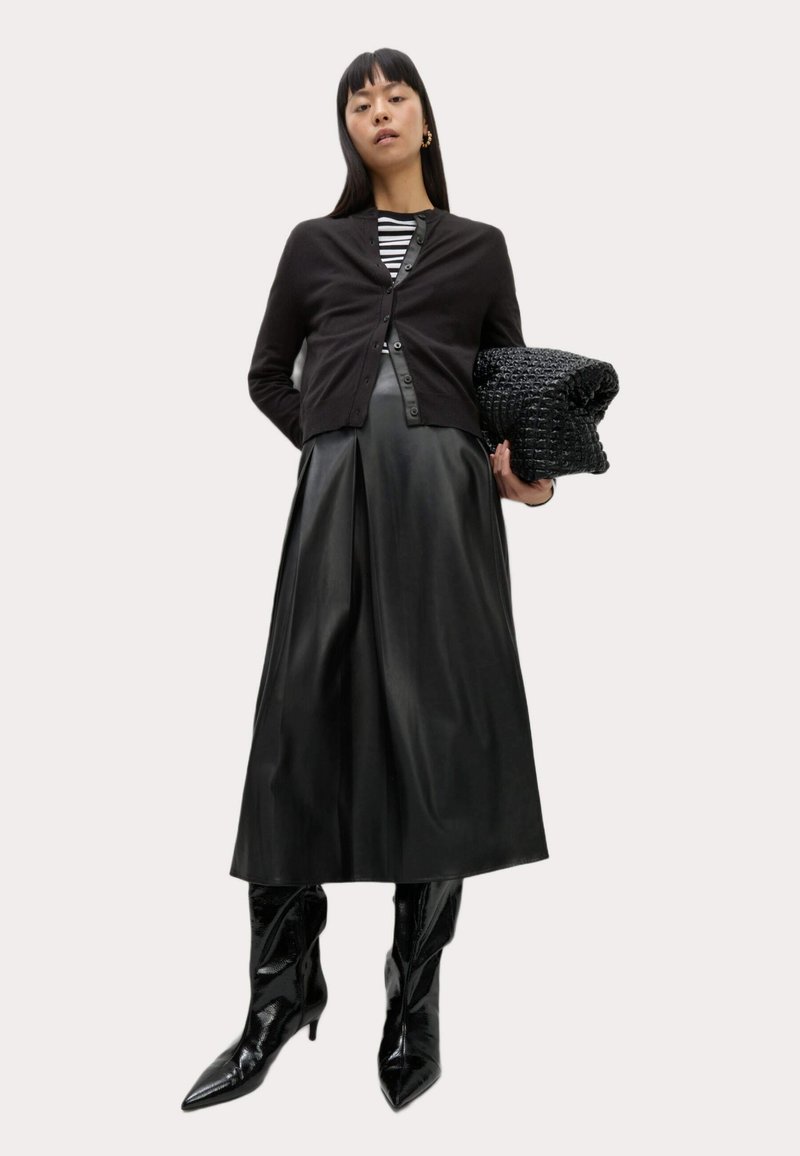 Un cardigan noir sur un top rayé, associé à une jupe plissée en cuir noir et des bottes à bout pointu noires et brillantes. Tenant un sac noir texturé.