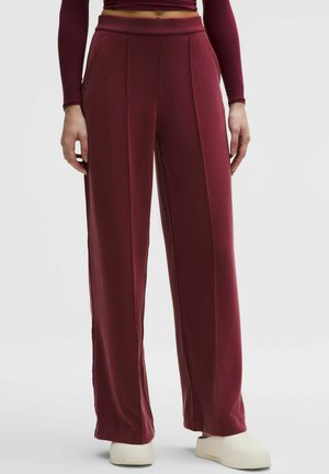 Person trägt weinrote hochgeschnittene, weit geschnittene Hose mit vorderen Nähten, kombiniert mit beigen Slip-On-Schuhen vor einfachem Hintergrund.