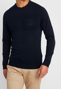 Granatowy sweter dzianinowy z długimi rękawami, ściągacze przy mankietach i hemie, z teksturowanym górnym pasem oraz wytłoczonym logo na klatce piersiowej.