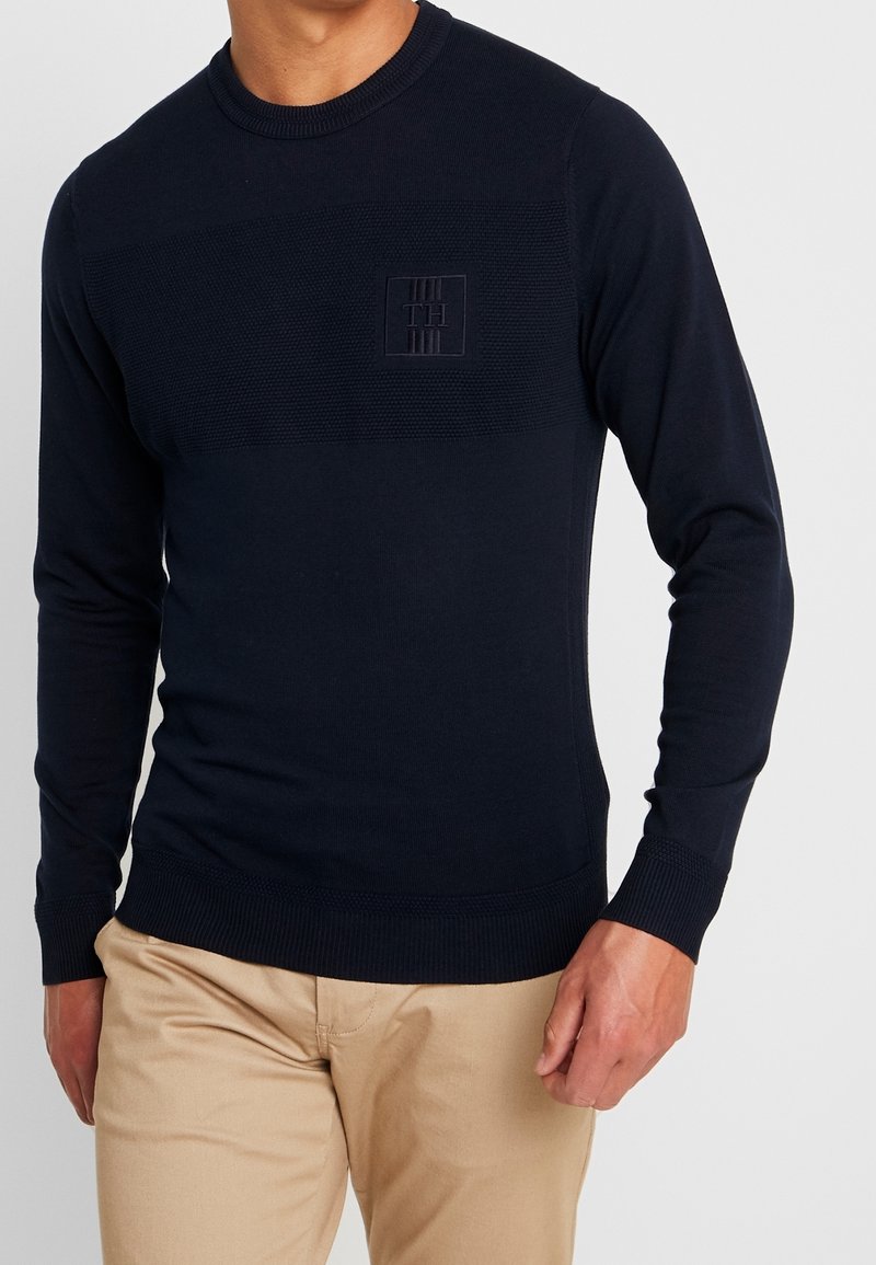 Marineblau gestrickter Pullover mit langen Ärmeln, gerippten Bündchen und Saum, ausgestattet mit einem strukturierten oberen Band und einem geprägten Logo auf der Brust.