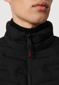 Chaqueta acolchada negra con cuello alto, cremallera frontal y tejido texturizado con un acabado suave. Acento del logo rojo cerca de la cremallera.