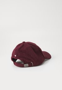 Casquette de baseball en velours côtelé bordeaux avec une visière courbée, une sangle ajustable et un petit détail brodé au dos indiquant '85'.