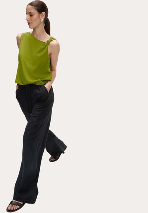 Top verde senza maniche con un dettaglio a nodo su una spalla, abbinato a pantaloni neri a gamba larga e sandali neri. La modella ha le mani nelle tasche.