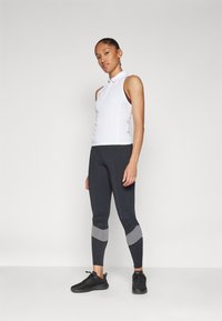 Polo bianco senza maniche con colletto, abbinato a leggings grigio scuro con dettagli grigi. Scarpe da ginnastica nere completano l'outfit.