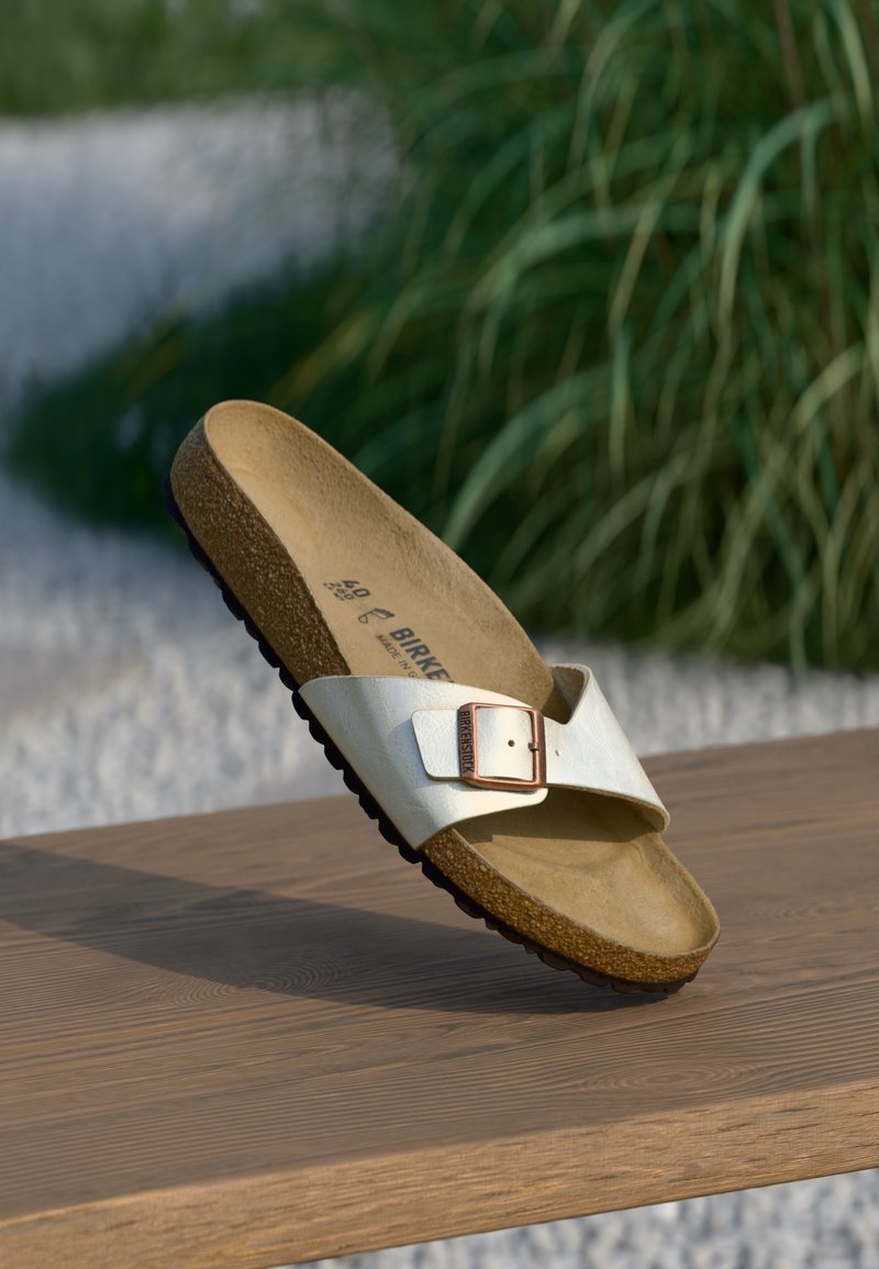 Sandale Birkenstock beige avec une seule bride à boucle réglable reposant sur une surface en bois à l'extérieur avec des plantes vertes en arrière-plan.