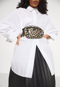 Borsetta a vita con stampa leopardata in tessuto nero, giallo e grigio; indossata sopra una camicia bianca oversize con bottoni e una gonna plissettata nera.