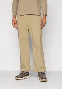 Beige, lightweight, water-resistant pants with a relaxed fit and elastic waistband. Worn with dark, rugged hiking shoes featuring beige accents.