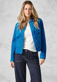 Blå bomullsjacka med knäppning, två framfickor och lång ärm, tillsammans med mörk denimjeans mot en texturerad bakgrund.