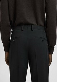 Widok z tyłu osoby noszącej czarne dopasowane spodnie z zapinanymi na guziki tylnymi kieszeniami oraz ciemnobrązowy sweter z długim rękawem.