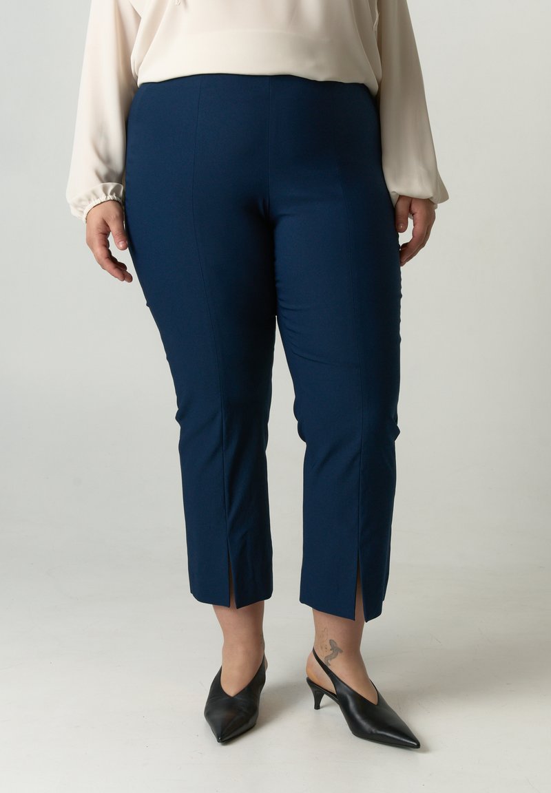 Navyblauwe tapered broek met zijsplits aan de zoom, gemaakt van een stretchstof. Draagt een lichtblouse met zwarte, puntige hakken.