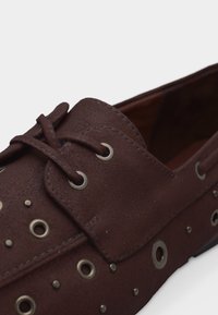 Καφέ σκούρο δερμάτινο μοκασίνι με δέσιμο κορδονιών και πολλαπλές μεταλλικές θηλιές και τρουκς που διακοσμούν το πάνω μέρος και τις πλευρές.