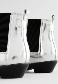 Botines metálicos plateados hasta el tobillo con paneles elásticos negros acanalados, tacones redondeados y tiradores en la parte posterior para un fácil calzado.