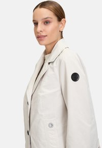 Light beige trench coat with a slightly shiny finish, black button accents, and a logo patch on the left sleeve. Standard collar and pockets.