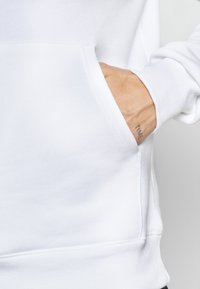 Sweatshirt blanc avec une poche kangourou, des poignets et un ourlet côtelés. Présente une texture de tissu douce et des détails de couture visibles.