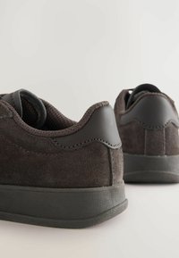 Ciemnobrązowe zamszowe sneakersy z teksturowanymi czarnymi podeszwami, zbliżenie na pięty na jasnym neutralnym tle.