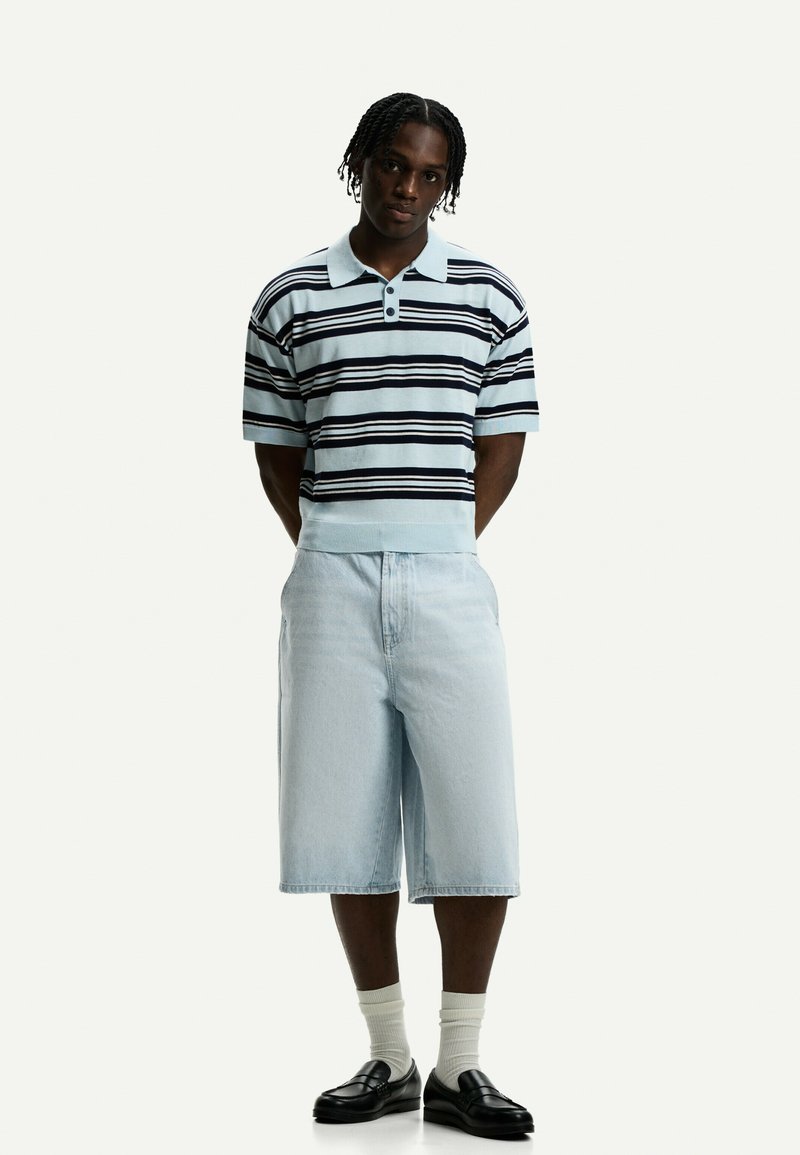 Polo a righe in azzurro chiaro e blu navy con due bottoni, abbinato a pantaloni shorts in denim azzurro chiaro e mocassini neri, completato con calze bianche.
