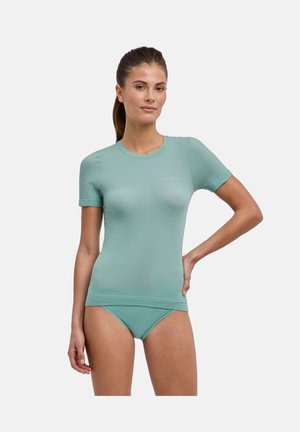 Femme aux cheveux bruns en queue de cheval portant un t-shirt moulant à manches courtes vert d'eau clair et un bas de bikini assorti sur un fond blanc uni.