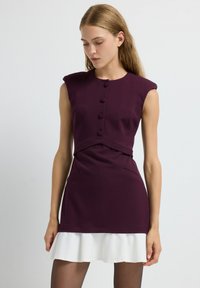 Robe à manches courtes de couleur violet foncé avec un corsage ajusté, des détails en boutons et un ourlet blanc à volants. Tissu doux au toucher lisse.