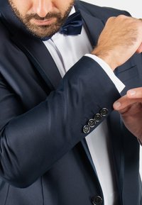 Veste de costume bleu marine avec finition satinée, accompagnée d'une chemise blanche et d'un nœud papillon. La veste présente des accents de boutons sur les poignets.