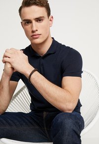 Un tricou polo bleumarin cu mâneci scurte, cu nasturi la guler, asociat cu o pereche de blugi negri. O brățară împletită împodobește încheietura.