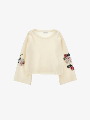 Kermanvärinen neule, jossa on pyöreä pääntie ja kellohihnat. Hihnoissa on vaaleanpunaisia ja punaisia kukkakirjontoja. Pehmeä rakenne.