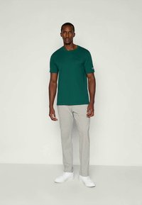 T-shirt verde a maniche corte con scollatura tonda, abbinato a pantaloni grigi chiari e testurizzati e sneakers bianche, in piedi contro uno sfondo neutro.