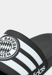 Μαύρη παντόφλα slide με υφασμάτινη λωρίδα που διαθέτει λευκή διαγώνια ρίγα και λογότυπο της FC Bayern Munich. Μαλακή σόλα.