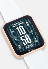 Reloj inteligente con una pantalla rectangular negra, acentos dorados, correa blanca y un fondo verde texturizado que muestra la hora y los datos de fitness.