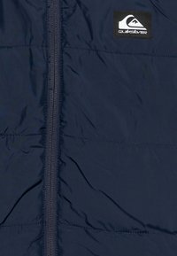 Mornarska puffer jakna s središnjim zatvaračem, pikastom teksturom i crnim Quiksilver logotipom na gornjem lijevom dijelu.
