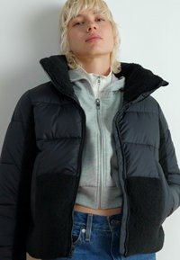 Dun svart med puffy design og svarte sherpa-detaljer, glidelås, høy krage, båret over en grå hettegenser, kombinert med blå jeans.