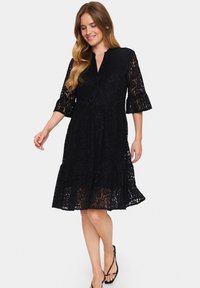Black lace dress with a v-neck, three-quarter flared sleeves, and a tiered skirt. Features intricate floral patterns and a semi-sheer texture.