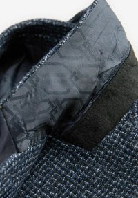 Sakoi prilagođen u tamnoplavom tvidu s teksturiranim uzorkom. Ima elegantan crni ovratnik i detaljno unutarnje podstavljanje s suptilnim printom.
