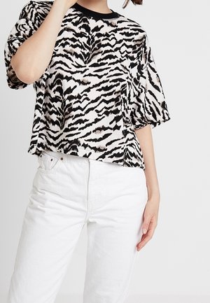 Vrouw draagt een zwart-witte tijgergestreepte top met korte mouwen en een witte hooggetailleerde spijkerbroek, staand tegen een effen achtergrond.