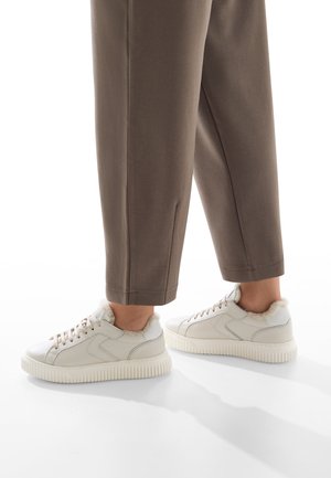 Persona che indossa pantaloni marroni e sneakers bianche con fodera in pile, in piedi su una superficie bianca.