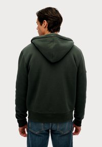 Donkergroene hoodie met een voorzak in kangoeroestijl, geribbelde manchetten en onderrand. Bevat een zachte, gestructureerde stof en een capuchon voor extra bescherming.