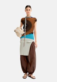 Wielokolorowy strój z plisowaną brązową bluzką, turkusowymi i zielonymi warstwowymi akcentami oraz luźnymi brązowymi spodniami. Trzyma miękką beżową torbę.