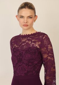 Top lungo a maniche con pizzo viola e motivi floreali, caratterizzato da un colletto scallop e una parte inferiore monocolore. Modella con capelli tirati indietro.