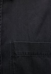 Gros plan sur un tissu en denim noir montrant une poche rectangulaire cousue et le bord de la couture du vêtement.