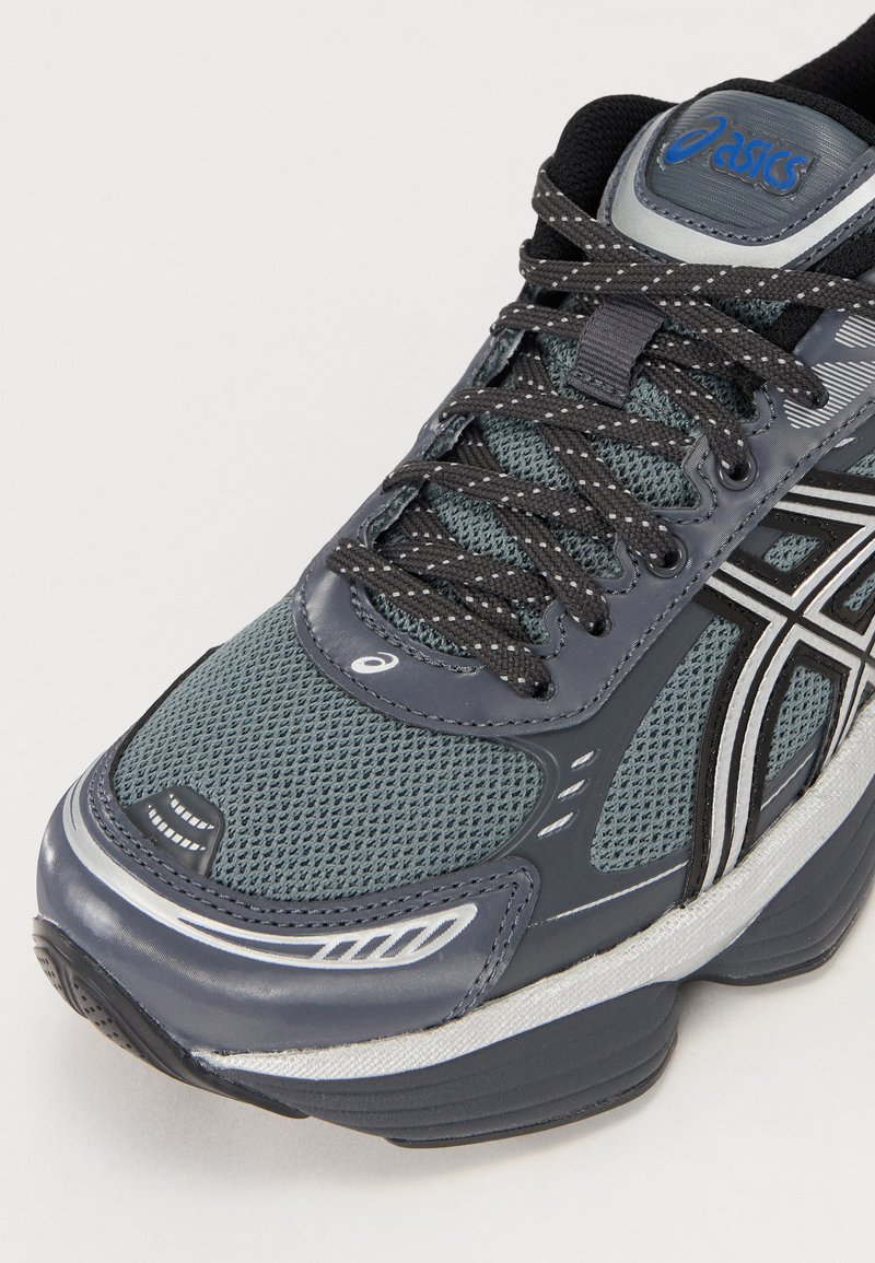 Zapatilla de running de malla gris con acentos negros, punta redondeada, suela gruesa, detalles reflectantes y cordones texturizados. Logo de Asics en la lengüeta.