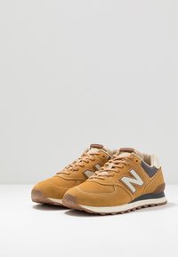 Hnědé semišové tenisky s béžovými akcenty a šedým síťovaným jazykem. Mají bílé logo "N" a texturovanou gumovou podrážku pro větší přilnavost.