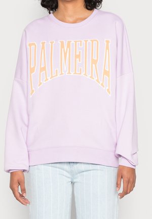 Osoba w lawendowej bluzie z dużym napisem „PALMEIRA” w kolorze brzoskwiniowym na piersi, w połączeniu z jasnoniebieskimi spodniami w paski.
