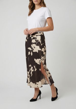 Mujer con camiseta blanca de manga corta, falda midi floral en negro y beige con una abertura lateral, y zapatos de tacón negros, de pie.